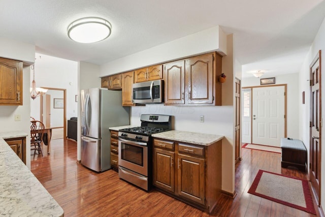 kitchen featuring an inviting chandelier, appliances with stainless steel finishes, brown cabinetry, dark wood-type flooring, and light stone countertops