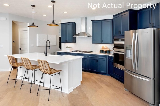 kitchen featuring light wood finished floors, blue cabinetry, appliances with stainless steel finishes, a kitchen bar, and wall chimney range hood