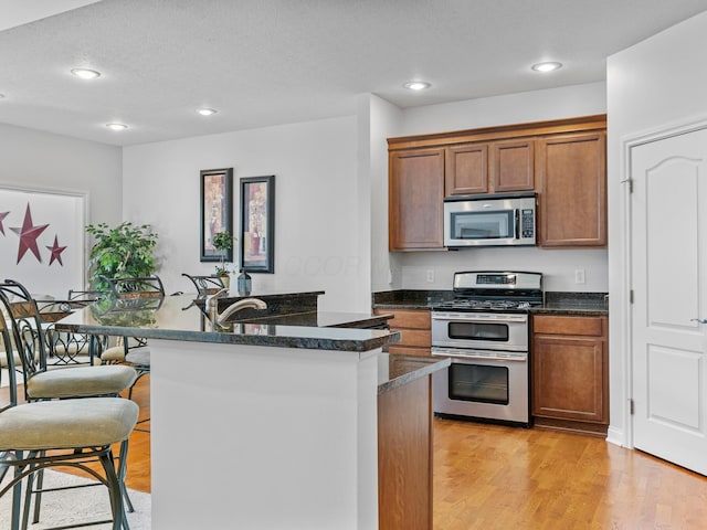 kitchen with recessed lighting, light wood-style flooring, appliances with stainless steel finishes, brown cabinets, and a kitchen island with sink