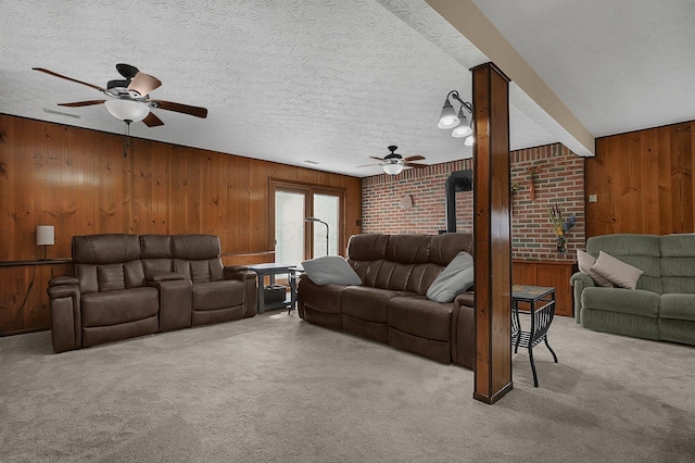 living room with carpet, a ceiling fan, a wood stove, wooden walls, and a textured ceiling