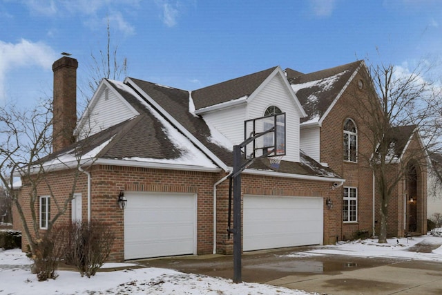 view of snowy exterior featuring a garage, brick siding, a chimney, and roof with shingles