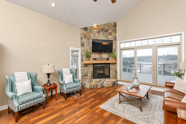 living area featuring high vaulted ceiling, a stone fireplace, wood finished floors, a ceiling fan, and baseboards