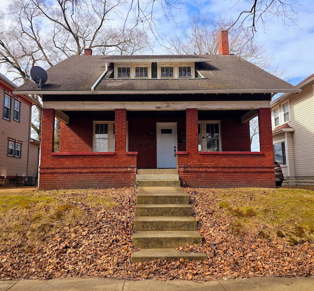 bungalow-style house featuring covered porch, roof with shingles, a chimney, and brick siding