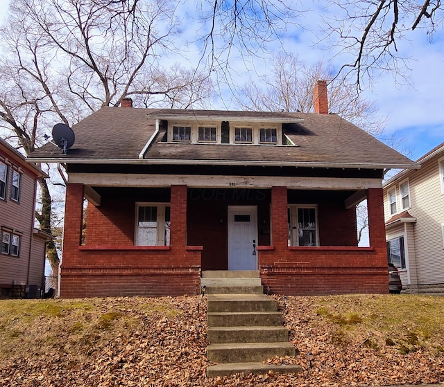 bungalow-style house with covered porch, a shingled roof, a chimney, and brick siding