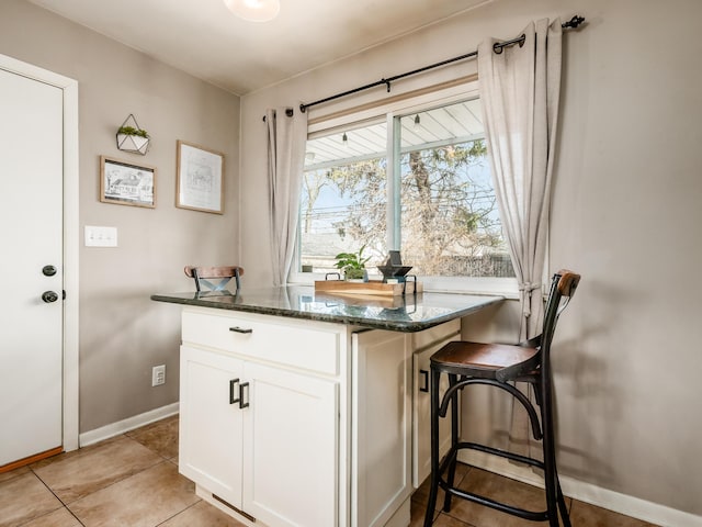 kitchen with a breakfast bar, light tile patterned floors, white cabinets, dark stone counters, and baseboards