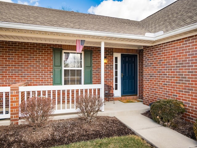 entrance to property with covered porch, brick siding, and roof with shingles