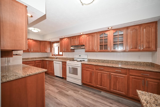 kitchen with light stone counters, brown cabinetry, a sink, white appliances, and under cabinet range hood