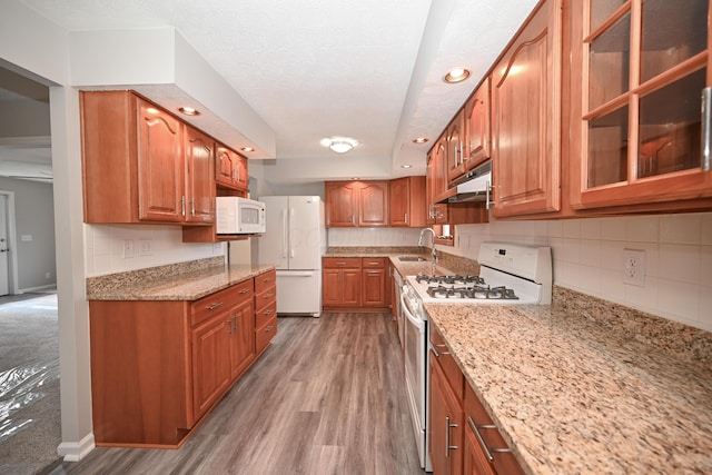 kitchen featuring light stone counters, under cabinet range hood, white appliances, a sink, and brown cabinets