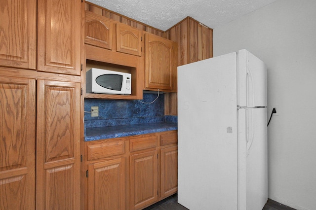 kitchen with dark countertops, white appliances, a textured ceiling, and brown cabinetry