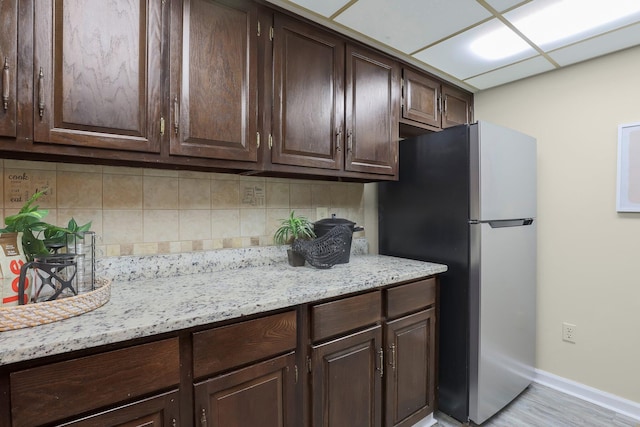 kitchen featuring dark brown cabinetry, baseboards, freestanding refrigerator, light stone countertops, and backsplash