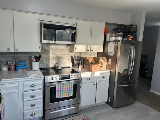kitchen featuring light wood-style floors, tile countertops, white cabinets, appliances with stainless steel finishes, and decorative backsplash