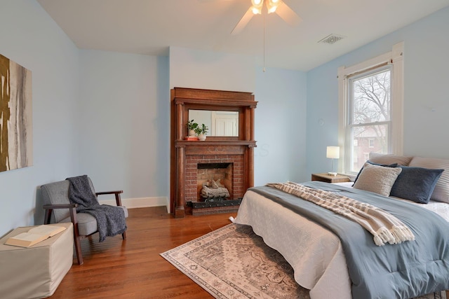 bedroom with ceiling fan, wood finished floors, visible vents, baseboards, and a brick fireplace