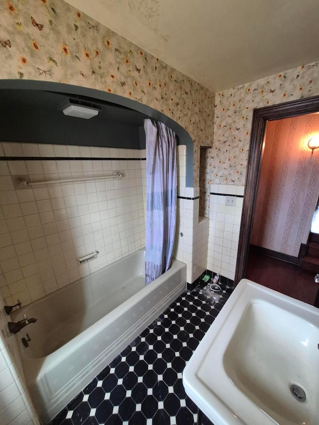 full bath featuring wallpapered walls, a wainscoted wall, tile patterned floors, shower / bathtub combination with curtain, and a sink