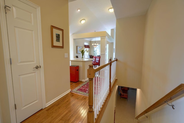 corridor with light wood finished floors, baseboards, lofted ceiling, an upstairs landing, and a notable chandelier