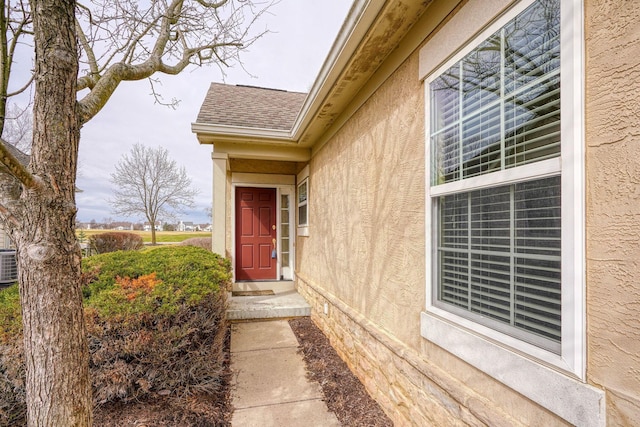 doorway to property featuring a shingled roof, central AC unit, and stucco siding