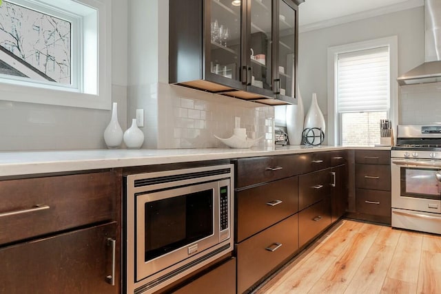 kitchen with stainless steel appliances, light countertops, light wood-style flooring, dark brown cabinets, and wall chimney exhaust hood