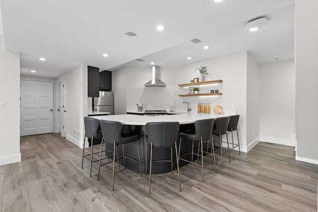 kitchen with a breakfast bar, a peninsula, freestanding refrigerator, light countertops, and wall chimney exhaust hood