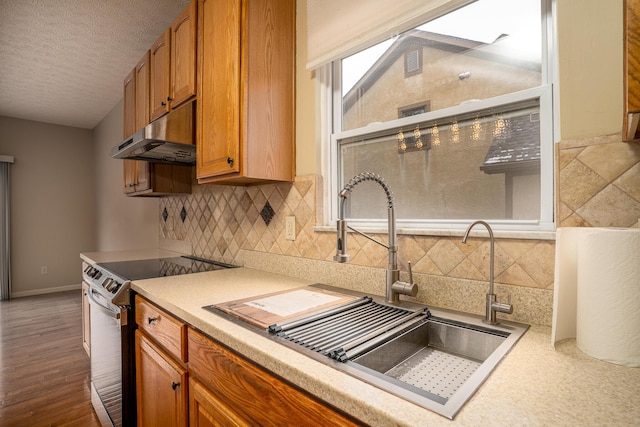 kitchen with a textured ceiling, under cabinet range hood, light countertops, stainless steel electric range, and brown cabinetry