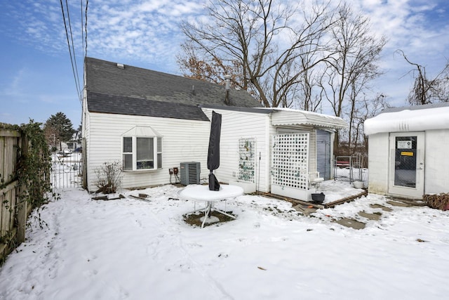 snow covered back of property with roof with shingles, fence, and central air condition unit