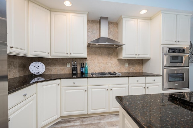 kitchen featuring stainless steel appliances, white cabinets, backsplash, dark stone counters, and wall chimney exhaust hood