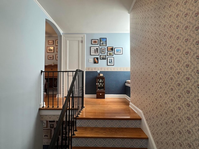 stairs featuring wallpapered walls, crown molding, wainscoting, wood finished floors, and arched walkways