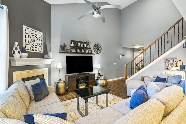 living room with a ceiling fan, a tiled fireplace, a towering ceiling, stairway, and wood finished floors