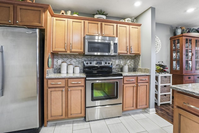 kitchen with brown cabinets, light stone countertops, stainless steel appliances, and backsplash