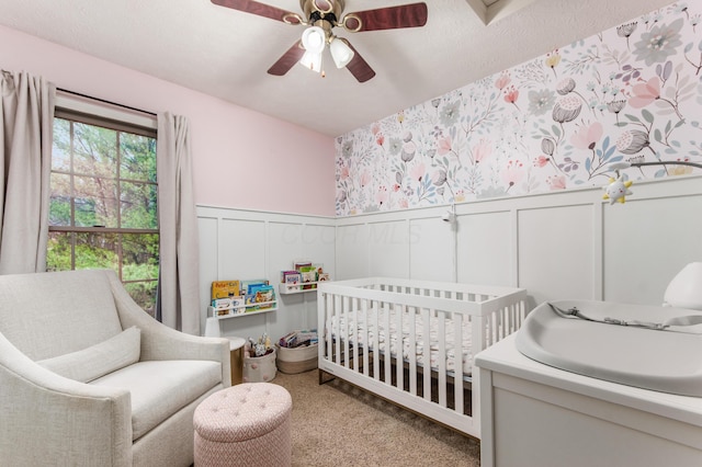 bedroom featuring a textured ceiling, carpet floors, wainscoting, a nursery area, and wallpapered walls