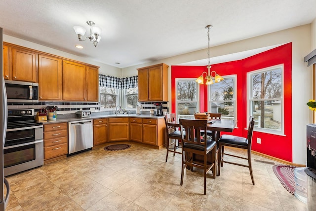 kitchen featuring stainless steel appliances, a sink, backsplash, brown cabinetry, and an inviting chandelier