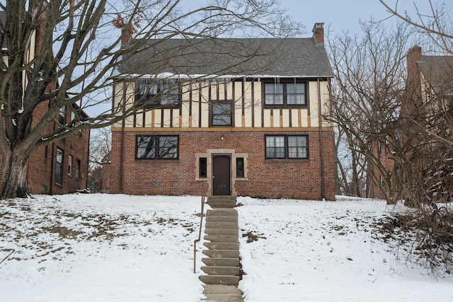 snow covered house featuring brick siding, stucco siding, a chimney, and roof with shingles