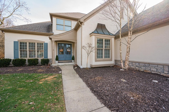 view of exterior entry featuring a shingled roof, a yard, stone siding, french doors, and stucco siding