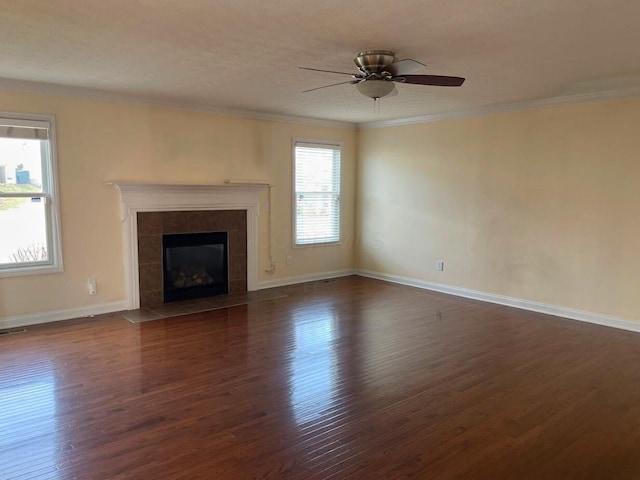 unfurnished living room featuring dark wood-style floors, a tiled fireplace, ornamental molding, ceiling fan, and baseboards