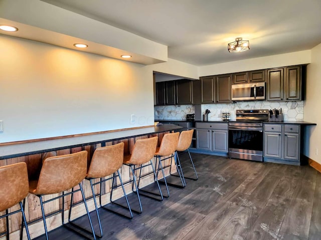 kitchen featuring decorative backsplash, dark countertops, appliances with stainless steel finishes, dark wood-type flooring, and a kitchen bar