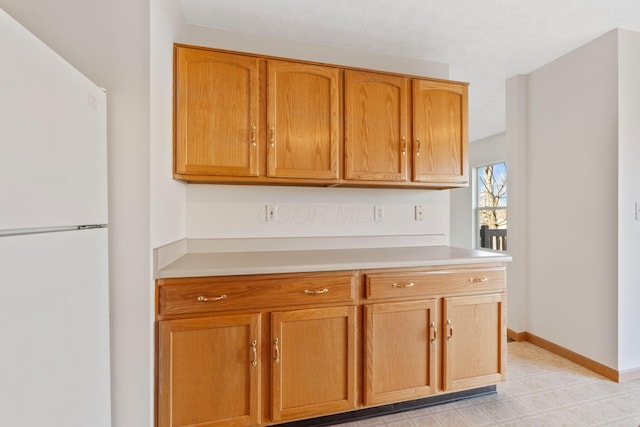 kitchen with brown cabinetry, freestanding refrigerator, light countertops, and baseboards