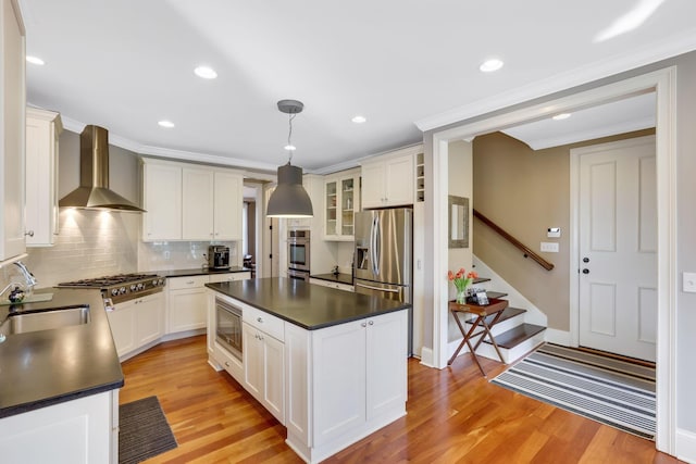 kitchen featuring light wood-style floors, dark countertops, appliances with stainless steel finishes, wall chimney range hood, and a sink