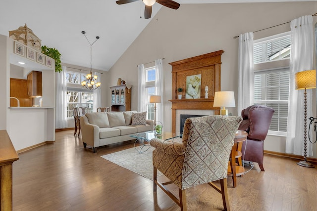 living area with baseboards, a tiled fireplace, wood finished floors, high vaulted ceiling, and ceiling fan with notable chandelier