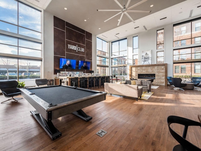 recreation room with a stone fireplace, a wall of windows, a towering ceiling, and hardwood / wood-style flooring