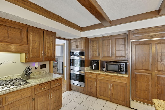 kitchen with light tile patterned floors, brown cabinetry, light stone counters, appliances with stainless steel finishes, and beamed ceiling