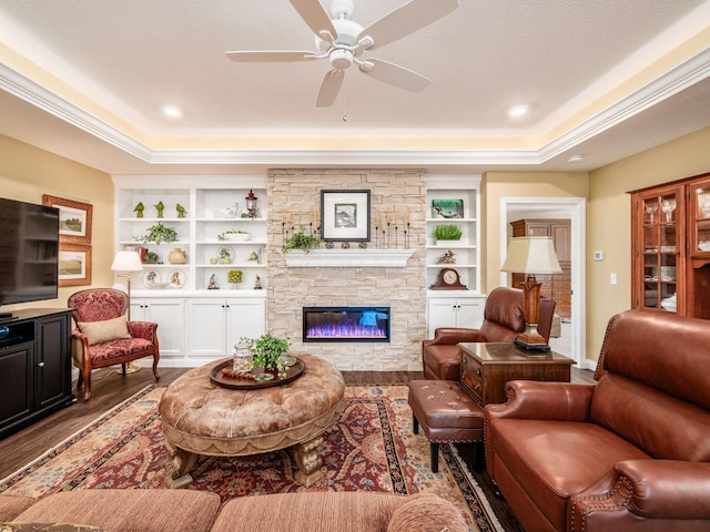 living area with built in shelves, wood finished floors, ceiling fan, a stone fireplace, and a raised ceiling