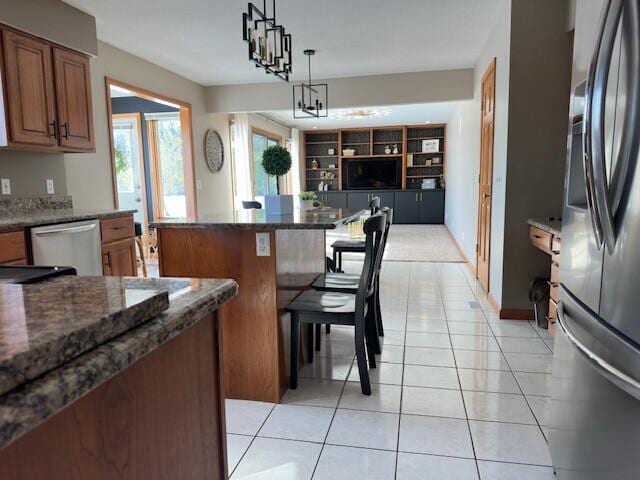 kitchen with pendant lighting, stainless steel appliances, brown cabinetry, light tile patterned flooring, and dark stone counters