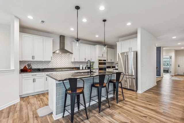 kitchen featuring wall chimney range hood, a sink, light wood-style floors, appliances with stainless steel finishes, and a kitchen breakfast bar