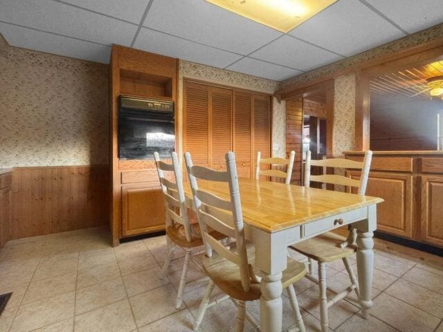 dining space featuring wallpapered walls, light tile patterned floors, a drop ceiling, and wainscoting