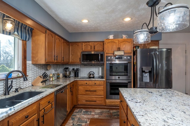 kitchen with brown cabinets, a sink, light stone counters, backsplash, and appliances with stainless steel finishes