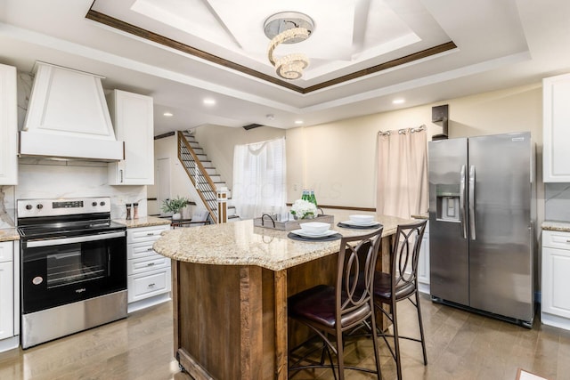 kitchen featuring light wood finished floors, a raised ceiling, appliances with stainless steel finishes, premium range hood, and white cabinetry