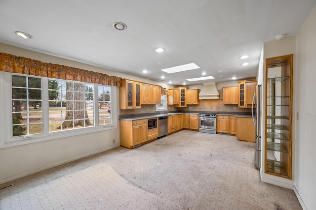 kitchen featuring custom range hood, dark countertops, stainless steel appliances, a skylight, and decorative backsplash