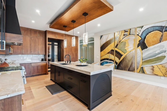 kitchen with light stone counters, custom exhaust hood, a kitchen island with sink, a sink, and light wood-type flooring