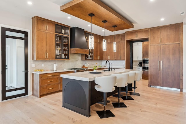 kitchen featuring premium range hood, light wood-type flooring, decorative backsplash, light countertops, and brown cabinets