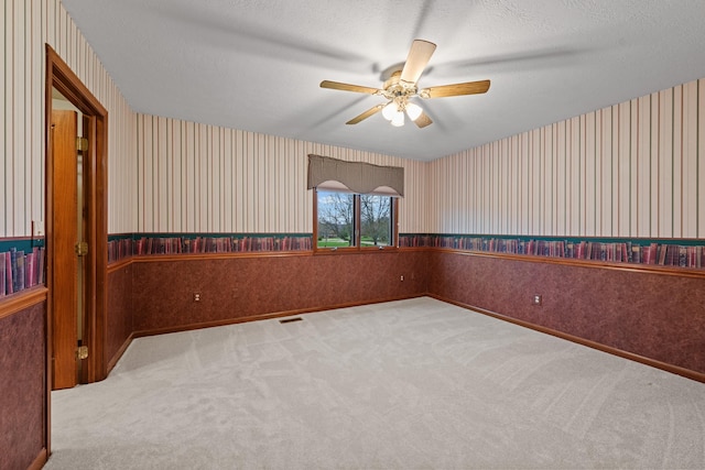 carpeted spare room featuring ceiling fan, a textured ceiling, visible vents, a wainscoted wall, and wallpapered walls