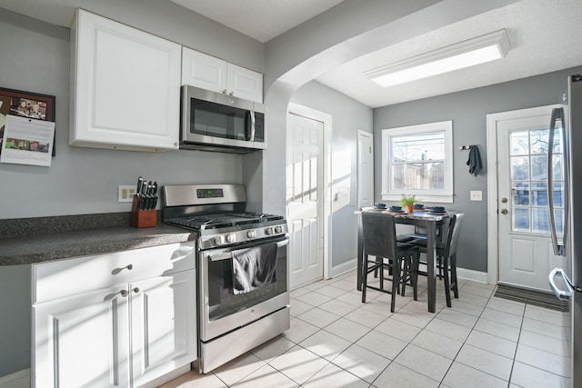 kitchen featuring light tile patterned floors, arched walkways, stainless steel appliances, white cabinets, and dark countertops