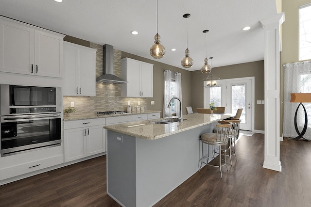 kitchen featuring dark wood-style flooring, a kitchen island with sink, a sink, appliances with stainless steel finishes, and wall chimney exhaust hood